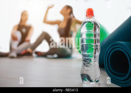 Support bouteille d'eau dans au tapis. Deux jeunes femmes s'asseoir derrière. L'un d'entre eux boivent de l'eau. Un autre regard sur son amie. Les modèles sont dans la salle de sport. Banque D'Images