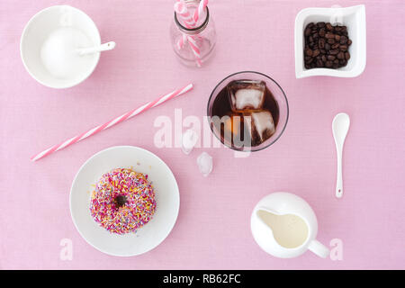 Dépoli rose donut coloré avec sprinkles, un verre rempli de préparer le café froid, le lait, le sucre, les grains de café et des pailles sur fond rose. Vue d'en haut. Banque D'Images