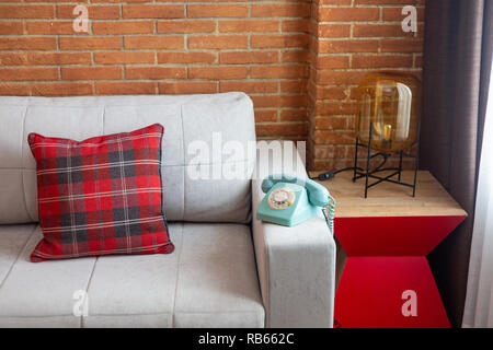 Téléphone vintage bleu sur le dessus d'un canapé gris avec un coussin tartan rouge, en plus d'une petite table avec lampe moderne. Mur de briques. Banque D'Images