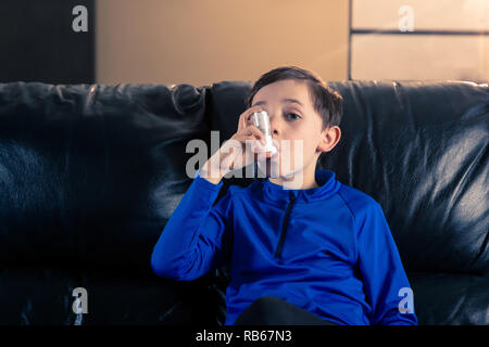 Garçon de 8 ans, portant des vêtements sportifs à l'aide d'un inhalateur d'asthme assis dans un canapé. Concepts : la santé, la maladie, le dopage Banque D'Images