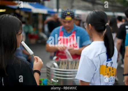 Une jeune femme est en train de manger un popsicle thaïlandais alors que son ami est en attente pour le sien en face du vendeur stand au marché de Chatuchak, le plus grand et le plus fa Banque D'Images