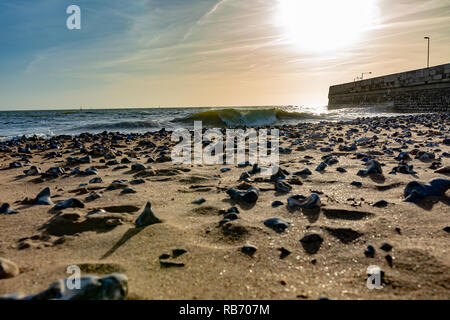 Photographie de paysage prises à partir de point de vue basse sur une plage de sable de stoney avec le fracas des vagues et mur du port en arrière-plan. Banque D'Images