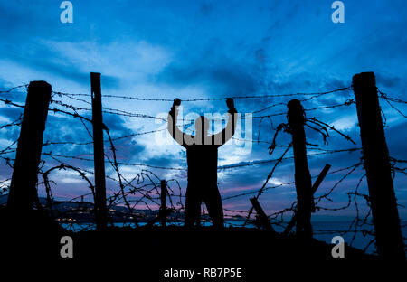 Silhouette of man looking through Barbed Wire/ le fil barbelé clôture à la nuit. Brexit, immigration, asile concept... Banque D'Images