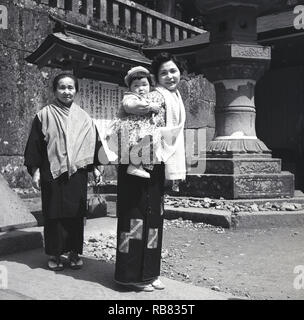 Années 1950, historiques, japonais mère, la fille et la visite d'un temple, les deux femmes portant des vêtements traditionnels, avec la mère portant son grand bébé sur son dos, le Japon. Banque D'Images