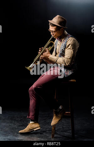 Mixed Race male élégant musicien jouant à la trompette en position assise sur une chaise sur noir Banque D'Images