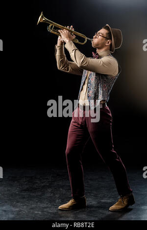 Young mixed race male jazzman dans hat jouer à la trompette sur noir Banque D'Images