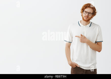 Portrait de mécontents et déçus drôle homme avec barbe rousse lunettes en fronçant les sourcils et plissant les yeux tout en regardant et pointant vers la gauche avec l'expression d'incertitude et n'aiment pas les créances douteuses Banque D'Images