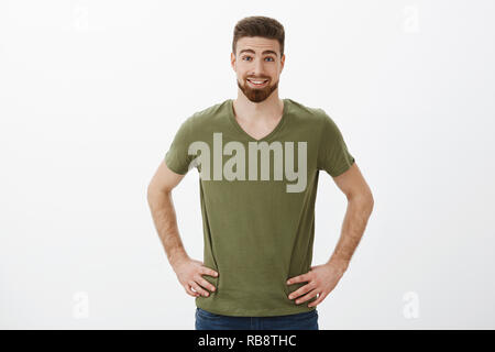 Surpris et enthousiaste de bon à barbu athlétique t-shirt en tenant les mains sur les hanches et le sourire de l'amusement et des sourcils étonnement détendue posant sur fond blanc Banque D'Images