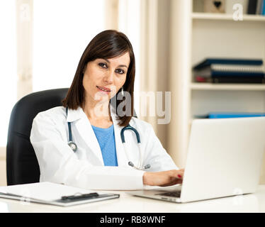 Portrait of happy jolie femme médecin en blouse blanche travail avec ordinateur portable et de l'écriture sur l'allégement assis sur clinic hospital office desk Banque D'Images
