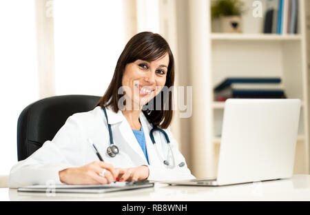 Portrait of happy jolie femme médecin en blouse blanche travail avec ordinateur portable et de l'écriture sur l'allégement assis sur clinic hospital office desk Banque D'Images