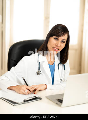Portrait of happy jolie femme médecin en blouse blanche travail avec ordinateur portable et de l'écriture sur l'allégement assis sur clinic hospital office desk Banque D'Images