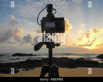 Appareil photo numérique reflex monté sur trépied photographier le lever du soleil sur l'océan. Ciel coloré, se concentrer sur l'appareil-photo, image à l'écran, l'arrière-plan flou. Banque D'Images