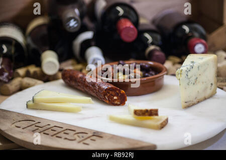 Présentation en gros plan d'une planche de fromages et charcuterie comprenant du fromage bleu, du fromage dur tranché, de la saucisse chorizo et un petit bol d'olives, posée contre un Banque D'Images