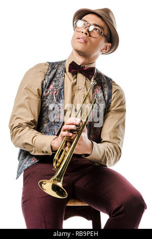 Pensive mixed race male jazzman holding trumpet et assis sur une chaise isolated on white Banque D'Images