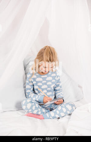 Cute kid en pyjama assis sur le lit avec les jambes croisées et l'écriture dans l'ordinateur portable Banque D'Images