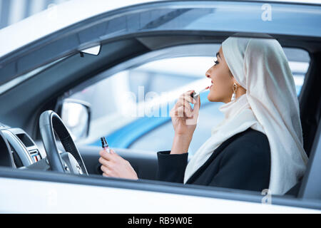 Jeune femme musulmane sitting in car et appliquant le maquillage Banque D'Images