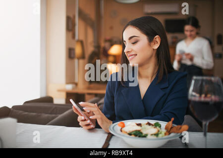 Cheerful young woman hold téléphone en mains et sourire. Elle a déjeuner au restaurant. Verre de vin rouge et de la salade reposer le bol sur la table Banque D'Images