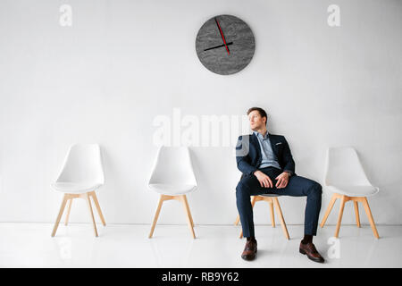 Young man in suit assis sur chaise blanche en salle blanche. Il s'appuyer au mur. Guy attendre fo vol en salle d'attente. Il siège seul Banque D'Images