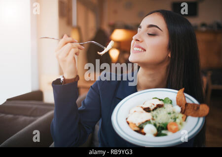 Jeune femme gaie du plaisir à manger de la salade. Elle sourit et maintenez-le morceau de fourchette. Model s'asseoir à table dans un restaurant Banque D'Images