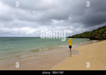 Jeune femme en veste jaune debout, marcher dans l'eau peu profonde sur une belle plage isolée, Ishigaki, Japon Banque D'Images