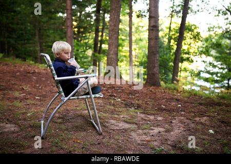 Un mignon petit Blond kid est boire du jus qu'il est assis dans une chaise de jardin vintage en camping près du lac. Banque D'Images