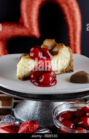 Morceau de gâteau au fromage gâteau cerise sur un piédestal avec des coeurs au chocolat ci-dessous et un bol de la sauce aux cerises. Fond noir avec cœur rouge. Banque D'Images