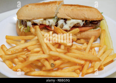 Philadelphia cheesesteak sandwich sur un sous bun. Couper en deux sur la plaque blanche avec frites en premier plan. Banque D'Images