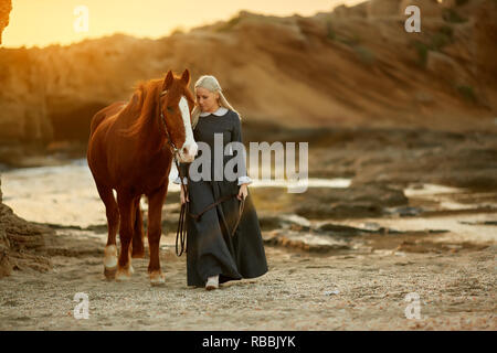 Dame mène un cheval au coucher du soleil le long de la mer dans un costume historique du 19e siècle Banque D'Images