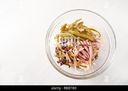 La préparation de la salade de saucisse (allemand wurstsalat) dans un bol en verre avec l'oignon, fromage et cornichons au vinaigre, fond clair avec copie espace, high angle view Banque D'Images