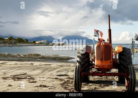 Un vieux tracteur vintage est situé sur une plage de sable trouver près d'une baie de la mer. Montagnes en arrière-plan et nuages de collecte avec une installation de pêche. Banque D'Images