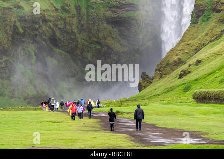 Skogafoss, Islande - 14 juin 2018 : Cascade sur falaise avec Green Grass Hills et de nombreux touristes qui marchent avec des parapluies et ponchos sur le sentier roa Banque D'Images