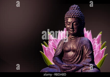 Statue de Bouddha assis en méditation poser contre fond sombre et fleur de lotus rose derrière. Banque D'Images