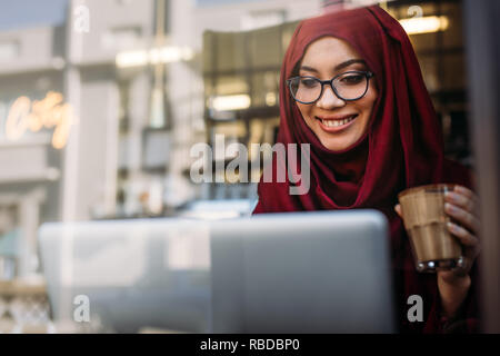 Smiling muslim woman working on laptop at coffee shop. Femme en hijab tenant une tasse de café et en regardant son portable dans la table à café. Banque D'Images