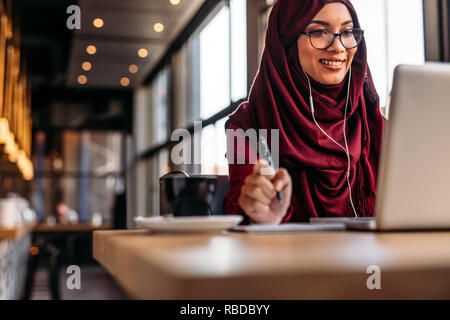 Belle femme en hijab au café après avoir vidéo conférence sur son portable. Femme musulmane avec stylo et bloc-notes ayant une vidéo sur son ordinateur portable à coff Banque D'Images