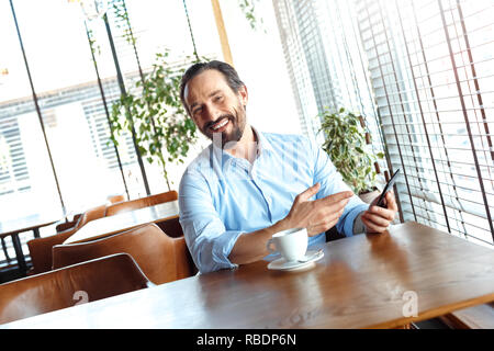 Commerçant homme mûr assis à table à café dans la journée avec tasse de café de l'écran montrant l'observation vidéo en ligne smartphone à rire caméra ch Banque D'Images