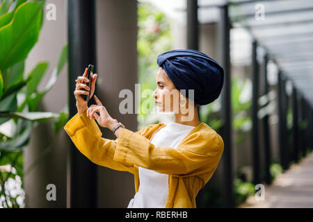 Une jeune et jolie femme Malais musulmans dans un élégant costume pastel et un turban prend une des selfies elle-même avec son smartphone sur la rue. Banque D'Images
