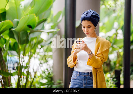 Une femme arabe du Moyen-Orient dans un foulard est SMS et messagerie instantanée sur son smartphone dans une rue de la ville pendant la journée. Banque D'Images