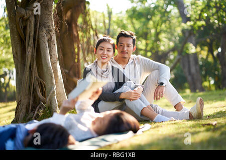 Deux enfants asiatiques little boy and girl having fun lying on grass lire un livre avec les parents assis à regarder en arrière-plan. Banque D'Images