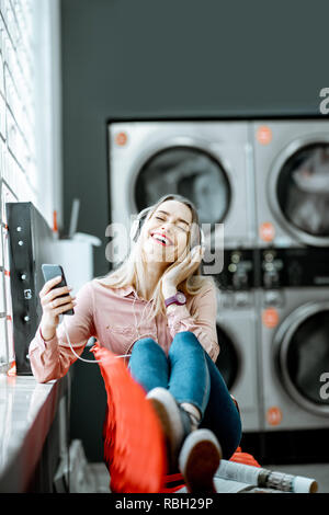 Jeune femme profiter de la musique en attente pour les vêtements à laver assis sur le fauteuil à la blanchisserie en libre-service Banque D'Images