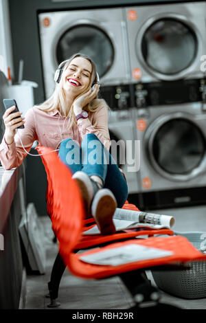 Jeune femme profiter de la musique en attente pour les vêtements à laver assis sur le fauteuil à la blanchisserie en libre-service Banque D'Images