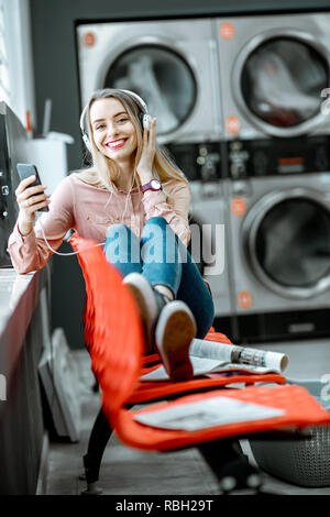 Jeune femme profiter de la musique en attente pour les vêtements à laver assis sur le fauteuil à la blanchisserie en libre-service Banque D'Images