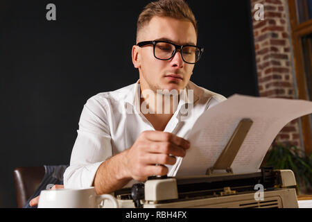 Auteur de livres portant des lunettes et une chemise blanche assise à la table avec machine à écrire et lire un nouveau chapitre de son livre Banque D'Images