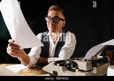 Auteur de livres portant des lunettes et une chemise blanche assise à la table avec machine à écrire et lire un nouveau chapitre de son livre Banque D'Images