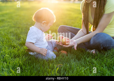 Belle jeune maman moderne jouant avec son mignon bébé fils comptant des pommes dans un parc ensoleillé. Le concept de l'éducation de l'enfant Banque D'Images