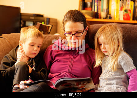 Une maman s'assit sur un canapé-lit un livre à ses deux jeunes enfants Banque D'Images