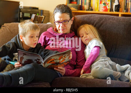 Une maman s'assit sur un canapé-lit un livre à ses deux jeunes enfants Banque D'Images