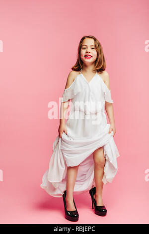 Jeune fille voulant être un adulte femme, portant robe et chaussures surdimensionnées sur fond rose. Fille de sa mère, looking at camera and smiling Banque D'Images