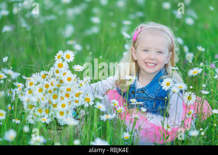 Cute little smiling baby girl en champ camomille petite blonde enfant avec couronne au chef de camomille portait un jean et la jupe rose bénéficiant d'hide-et-s Banque D'Images