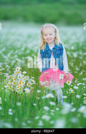 Cute little smiling baby girl en champ camomille petite blonde enfant avec couronne au chef de camomille portait un jean et la jupe rose bénéficiant d'hide-et-s Banque D'Images