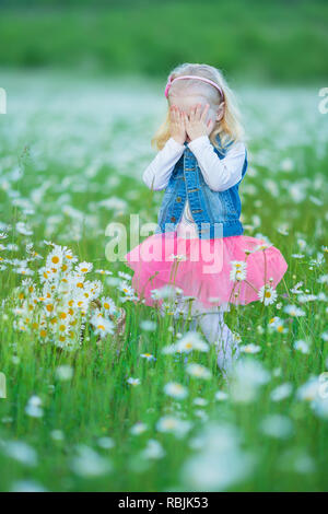 Cute little smiling baby girl en champ camomille petite blonde enfant avec couronne au chef de camomille portait un jean et la jupe rose bénéficiant d'hide-et-s Banque D'Images